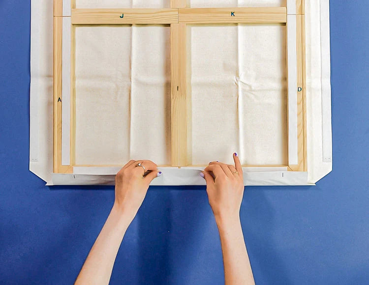 Hands assembling a wooden stretcher frame with adhesive strips on a blue surface for a Sculpd Canvas Painting Kit 