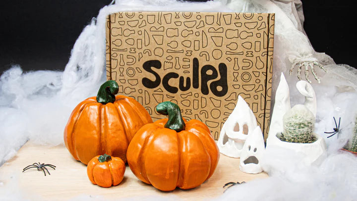 A Sculpd Pottery Kit box surrounded by handmade ceramic pumpkins, ghost decorations, and Halloween-themed props with cobwebs and spiders