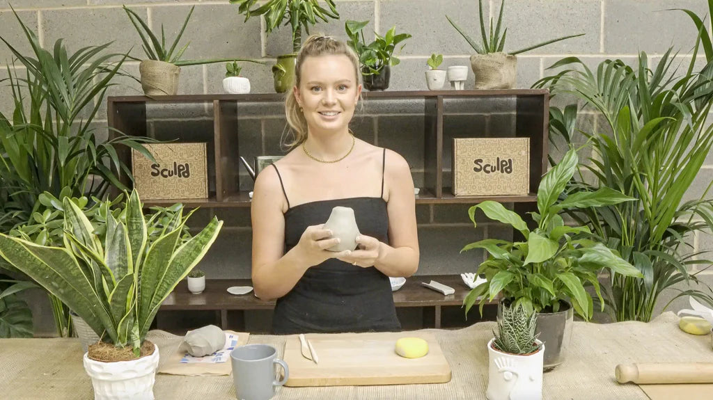 Smiling woman demonstrating pottery techniques, with Sculpd boxes and plants in the background