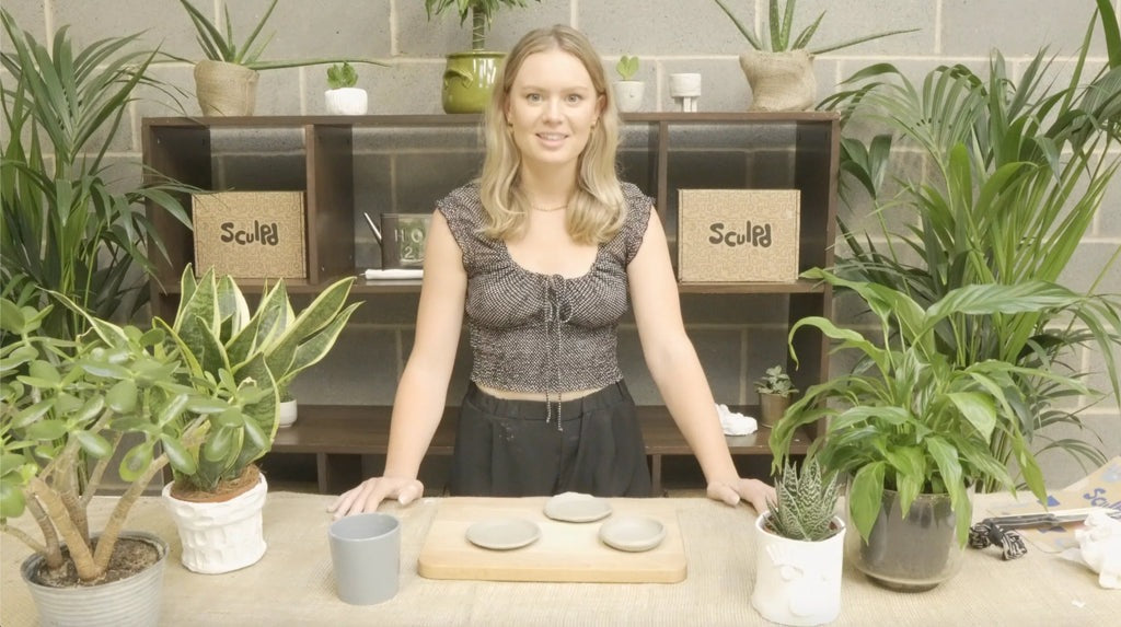 Smiling woman demonstrating pottery techniques, with Sculpd boxes and plants in the background